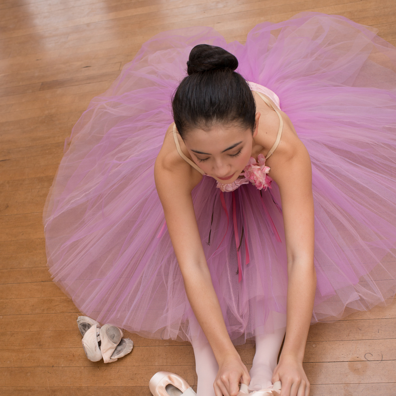 A teenage dancer in her tutu sits on the floor while putting on her pointe shoes.